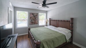 Een slaapkamer met een houten bed, een plafondventilator, twee ramen met zonwering en een schilderij aan de muur.