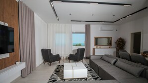 A modern living room with a sectional sofa, a white coffee table, and a flat-screen TV mounted on a wooden panel. There are two armchairs, a small round table, and a large window with curtains. - Wake Up to Lake Views – Cozy Comfort at Era e Liqenit” (Shkodër)
