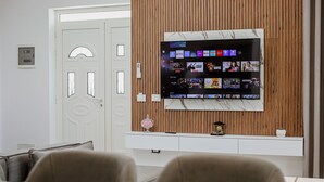 A modern office space with a large flat-screen TV mounted on a wooden wall, a white door with glass panels, and a white desk with a computer and a decorative item. - Wake Up to Lake Views – Cozy Comfort at Era e Liqenit” (Shkodër)