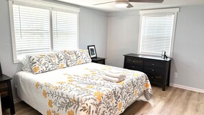 Quarto com cama, mesas de cabeceira, cômoda e duas janelas com persianas.