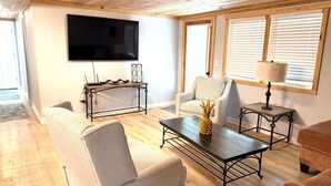 Uma sala de estar com televisão de tela plana, mesa de centro de madeira, poltrona estampada e uma mesinha com abajur.
