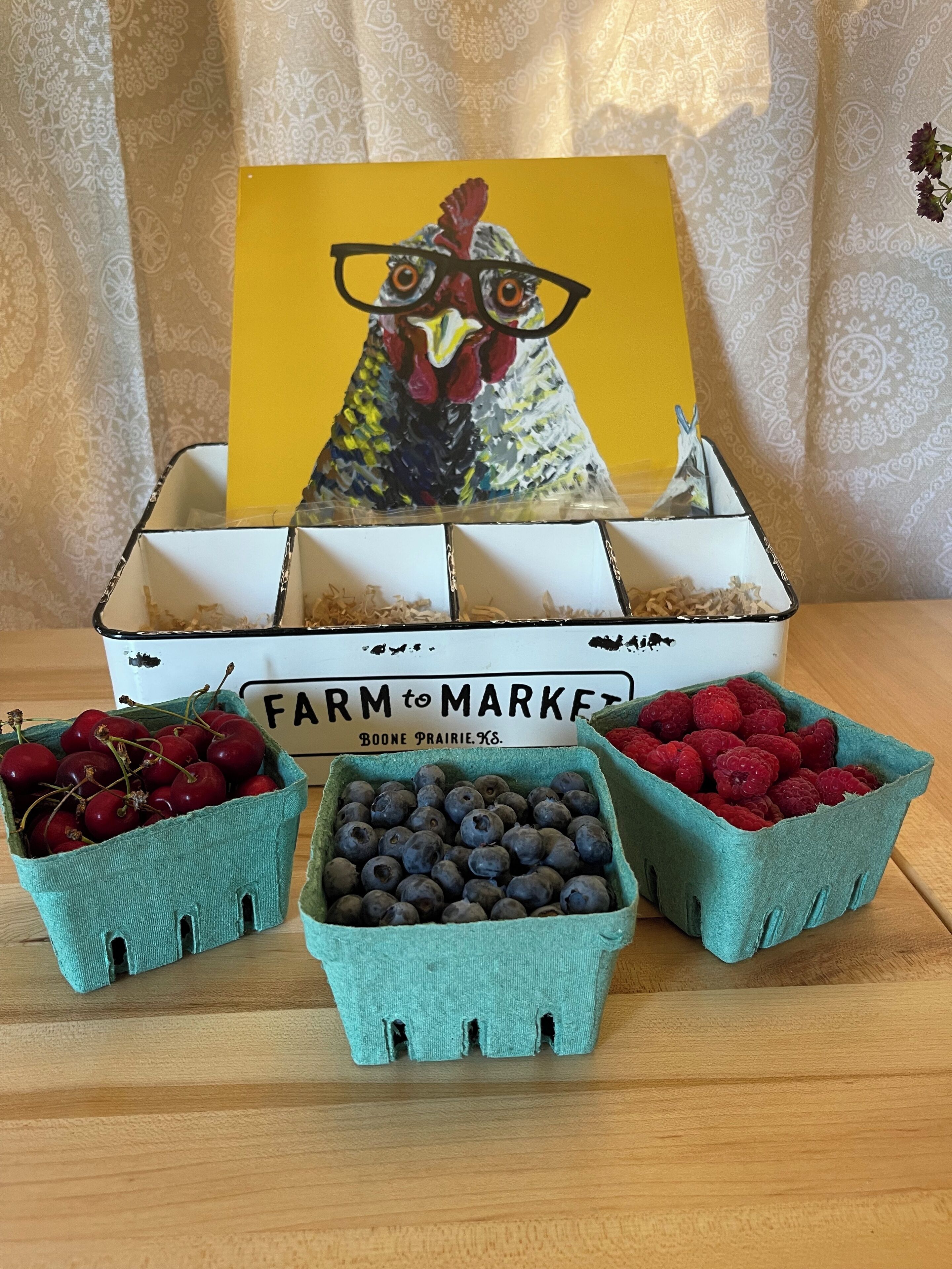 Een tafel met een 'Farm to Market' bord, een geschilderde haan en drie manden met kersen, bosbessen en frambozen.