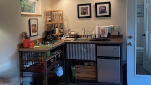 A compact kitchen with a built-in dishwasher, a coffee maker, and a small refrigerator. There are two framed pictures on the wall, a window with a view of greenery, and a shelf with kitchen utensils. - 3 Rivers Farm Guest Cottage. Charming Farm Stay cottage in Deming, Wa, (Deming)