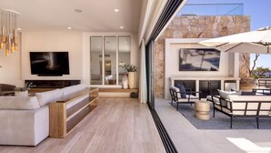 A modern living room with a flat-screen TV, a sofa, a coffee table, and a fireplace area with outdoor seating. - 4BR Ocean/Golf View • Pool & Transfers (Cabo San Lucas)