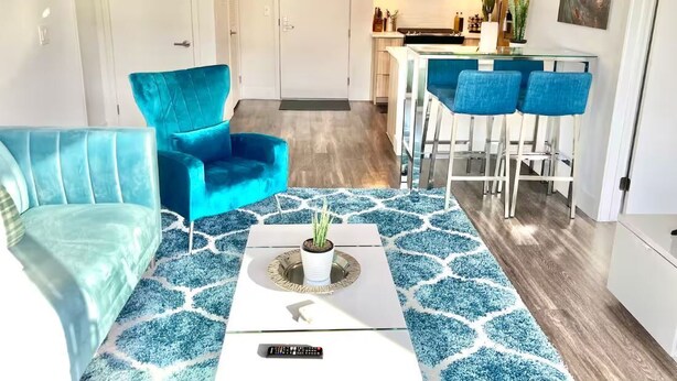 Een moderne woonkamer met een blauwgroene bank, een salontafel met een plant en een keukenhoek met een bar en een magnetron.