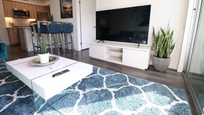 Een moderne woonkamer met een grote flatscreen televisie, een witte salontafel en een blauw gestreept vloerkleed.