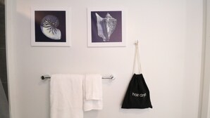 Een badkamer met een handdoekhouder waarop een witte handdoek en een zwarte tas met de tekst 'föhn' staan. Boven de handdoekhouder hangen twee ingelijste foto’s van schelpen aan de muur.