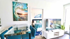 Een moderne woonkamer met een glazen eettafel, blauwe stoelen, een flatscreen televisie en kunstwerken aan de muren.
