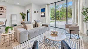 A modern living room with a grey sofa, a round coffee table, and a view of the outdoors through large windows.