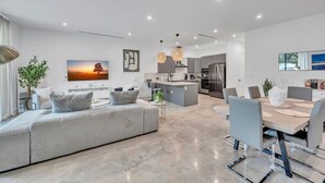 A modern living room with a grey sofa, a dining table with chairs, a kitchen area with a fridge and stove, and a TV mounted on the wall.