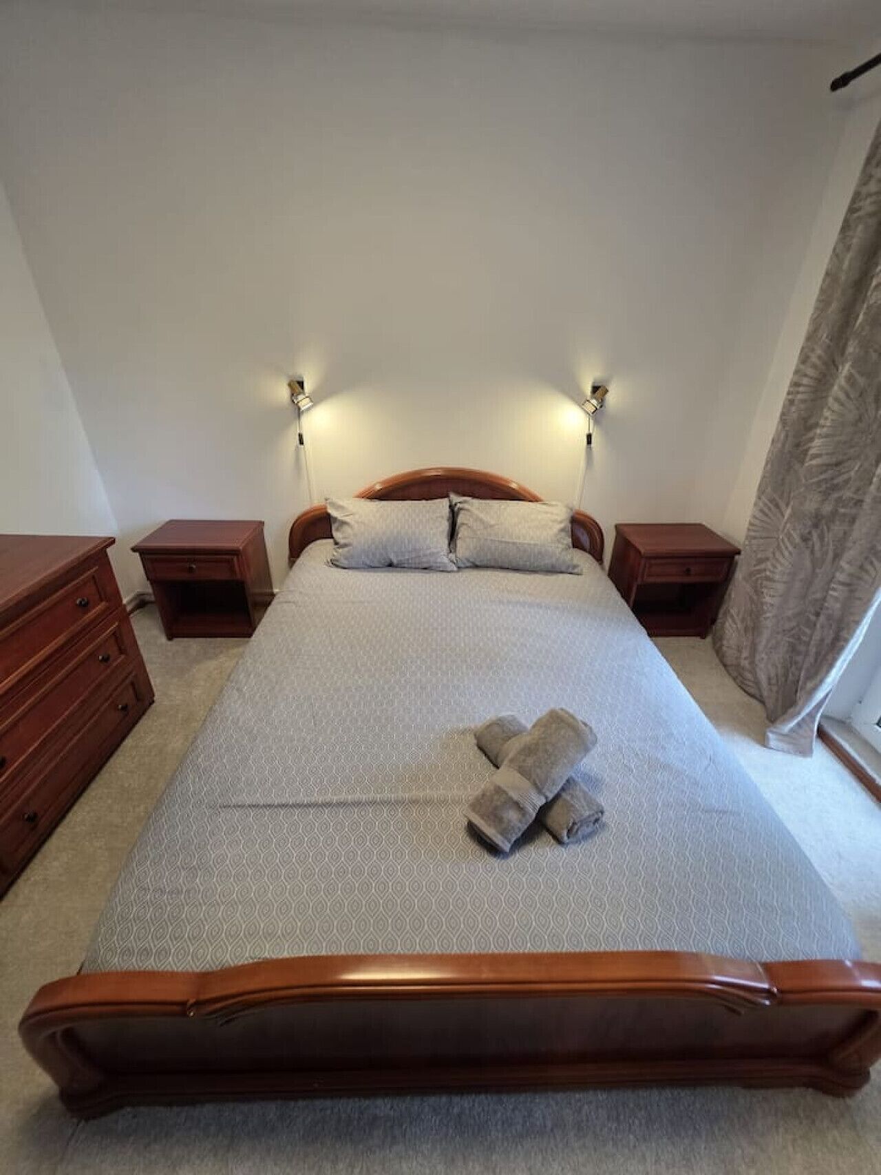 Una cama bien hecha con estructura de madera, dos mesitas de noche y dos lámparas de pared.