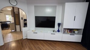 A modern living room with a large flat-screen TV, wooden flooring, and a white entertainment unit with books and decorative items. - Oradea Gateway | 3BR by River | Quick Airport Exit (Oradea)