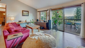 Un salon spacieux avec un canapé rouge, une table basse en bois et un grand pouf beige. Une porte-fenêtre coulissante donne accès à une terrasse extérieure équipée d’une table et de chaises.