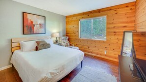 Une chambre avec un plafond en bois, un lit avec une literie blanche, une table de chevet avec une lampe, une chaise et une fenêtre donnant sur des arbres.