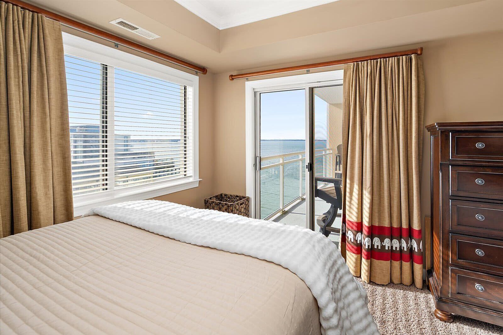 A hotel room with a bed, a wooden dresser, a window with blinds, and a view of the sea.