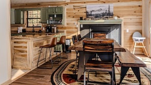 A rustic kitchen with wooden cabinets, a dining area with a long wooden table and chairs, and a fireplace. - Smoky Views & Game 4 BR Cabin w Hot Tub Near Wears Valley! (Sevierville)