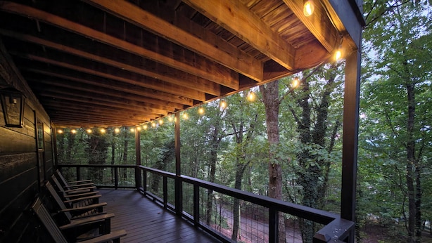 A wooden deck with string lights, benches, and a view of a forested area. - Smoky Views & Game 4 BR Cabin w Hot Tub Near Wears Valley! (Sevierville)