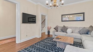 A living room with a grey sofa, a wooden coffee table, a black and white patterned rug, a TV mounted on the wall, and a chandelier. - The Main Stay - Steps from Ohio River, Shopping, & Dining on Main St (Madison)