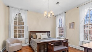 A bedroom with a wooden bed, a chair, a desk, and large windows with white curtains. - The Main Stay - Steps from Ohio River, Shopping, & Dining on Main St (Madison)
