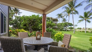 A covered outdoor seating area with wicker furniture and a round table, surrounded by lush greenery and palm trees. - Waikoloa Beach Villas K2 - Golf Course Front, Walk to Beach (Waikoloa)