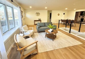 A spacious living room with a gray sofa, a wooden coffee table, and a white armchair. A large window provides a view of the outside.
