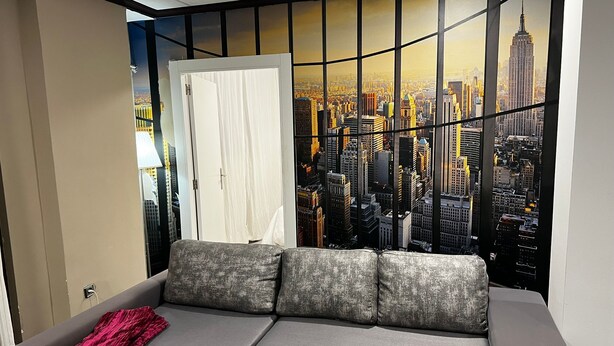 Ein Zimmer mit einem grauen Sofa, einem roten Kissen und einem großen Fenster mit Blick auf die Stadt.