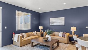 Ein Wohnzimmer mit zwei Sofas, einem Couchtisch und einem Esstisch mit Stühlen. Es gibt ein Fenster mit Jalousien, einen Feuerlöscher und ein Bild an der Wand.