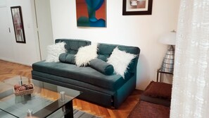 Een woonkamer met een donkergroene bank, witte, zachte kussens, een glazen salontafel met een rieten mand, en een moderne lamp.
