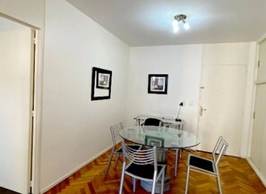 Une petite pièce bien éclairée, avec une table en verre et quatre chaises, des murs blancs et un plancher en bois.