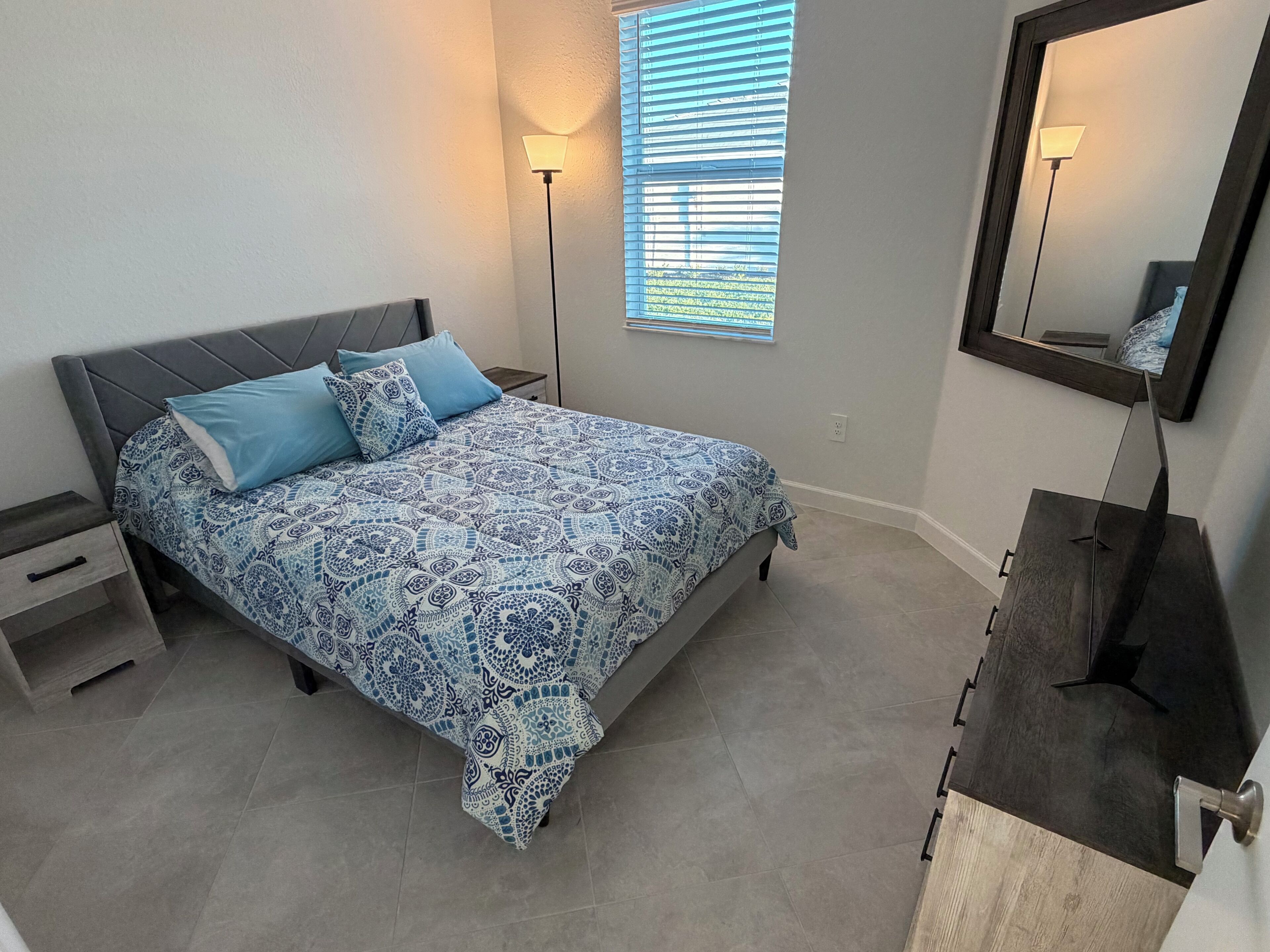 Un dormitorio con cama, mesitas de noche, cómoda, espejo y ventana con persianas.