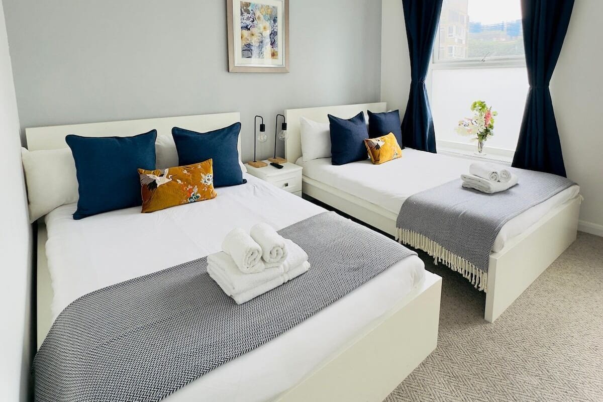 A hotel room with two beds, a bedside table with a lamp, a framed picture on the wall, and a window with curtains.
