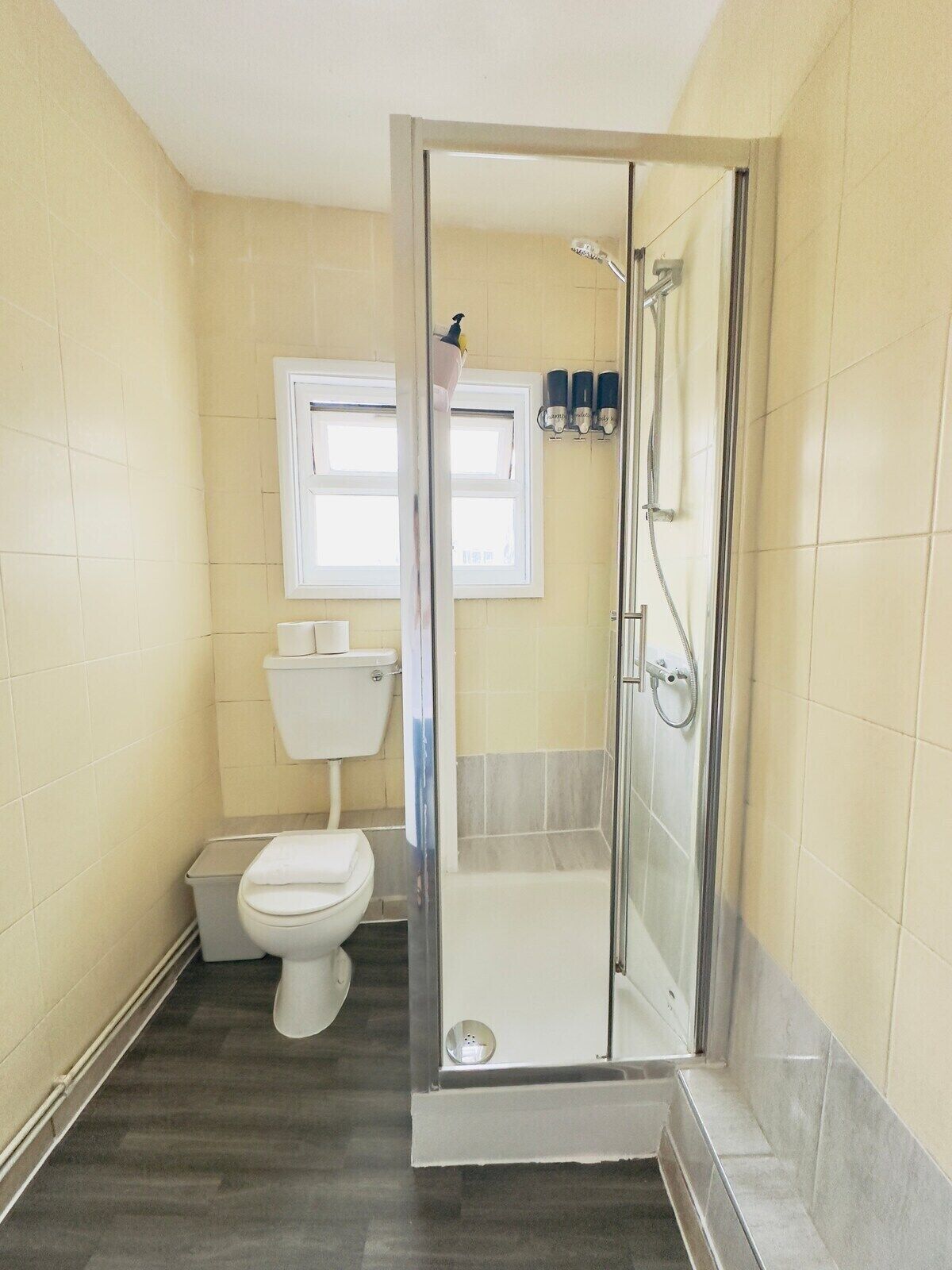 Un baño con inodoro, ducha con puerta de cristal y ventana sobre el inodoro.
