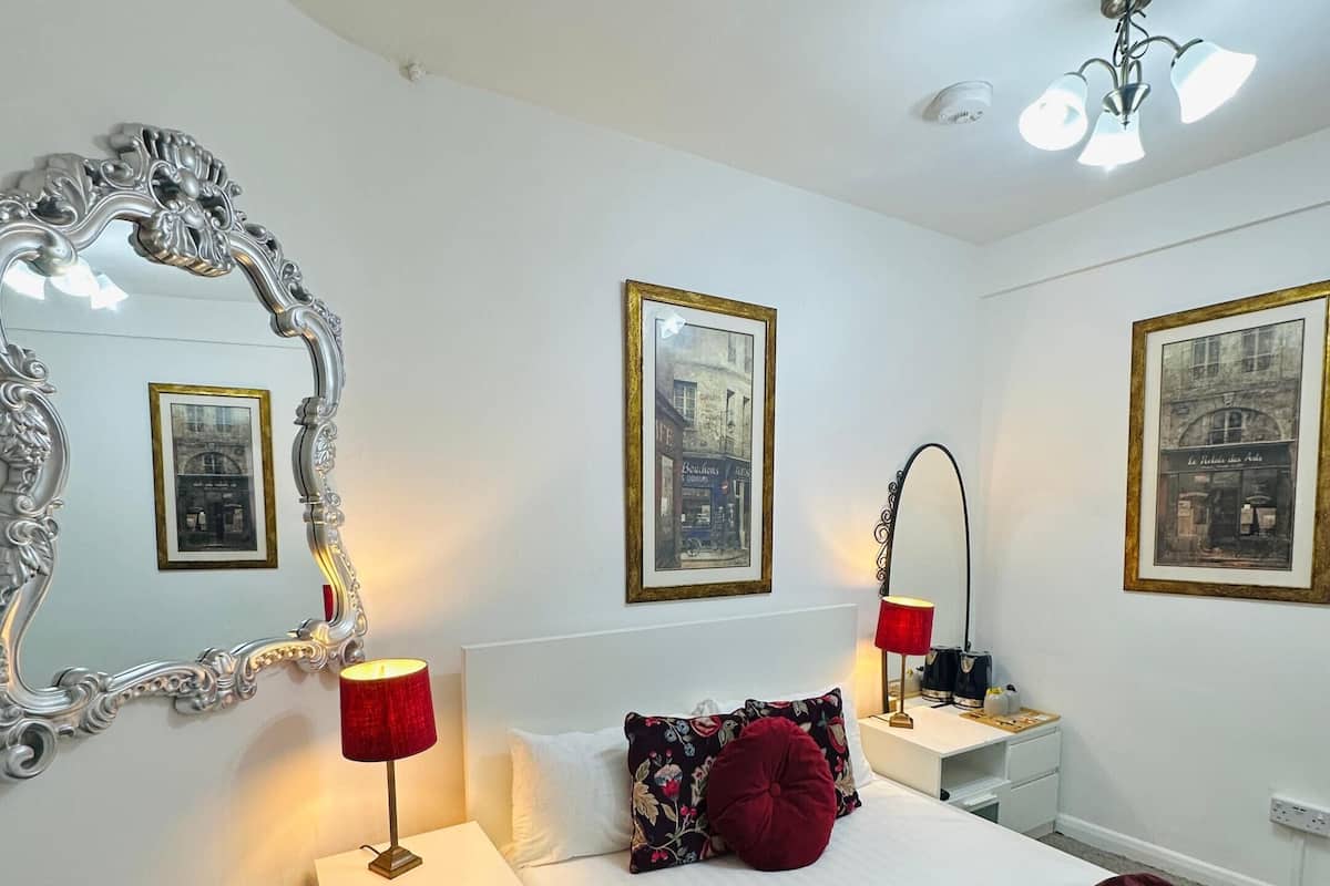 A bedroom with a large ornate mirror, a bed with a red blanket, two bedside tables with lamps, and framed pictures on the walls.