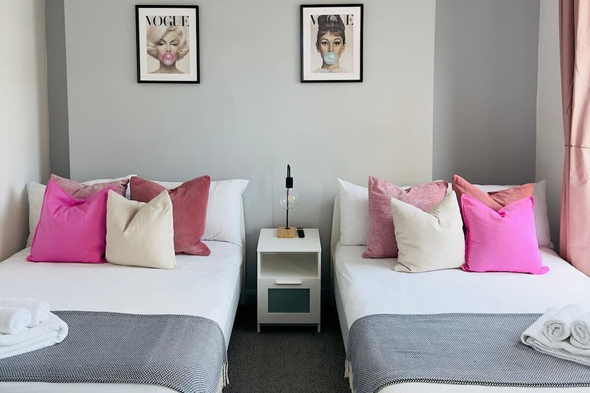A hotel room with two beds, a bedside table with a lamp, and two framed pictures on the wall.