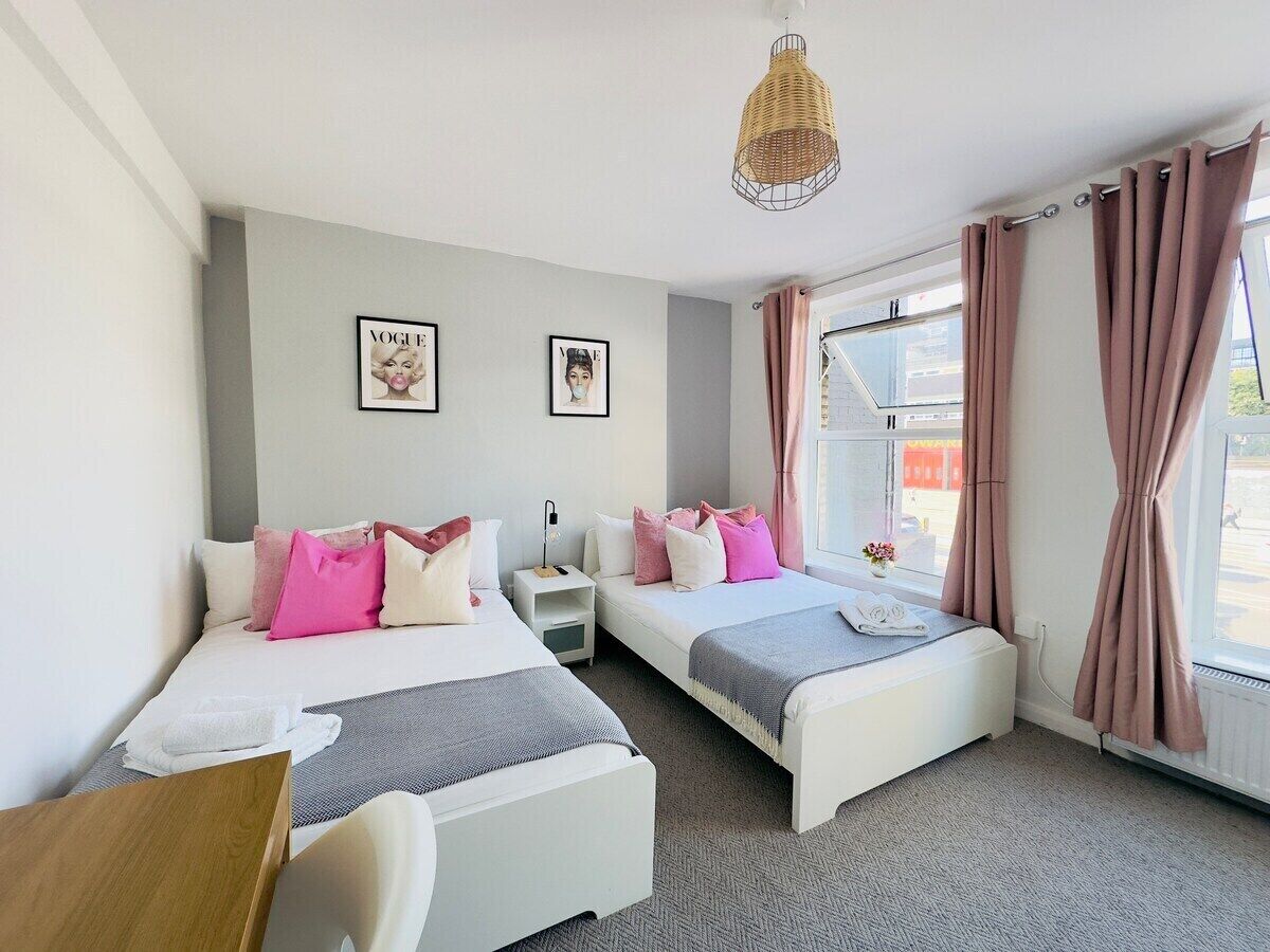 A hotel room with two beds, a desk, a chair, a small table, a nightstand, a lamp, and a window with curtains.