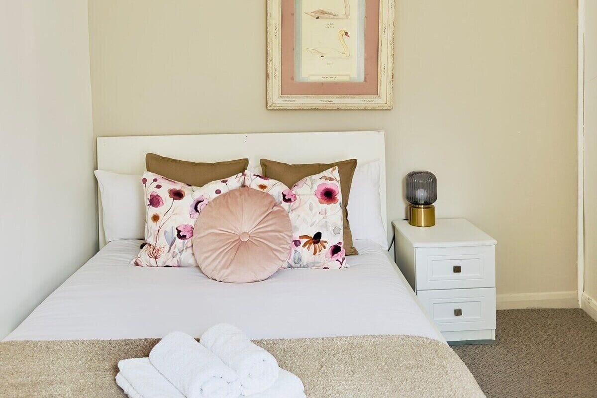 A neatly made bed with floral pillows, a framed picture on the wall, and a bedside table with a lamp.