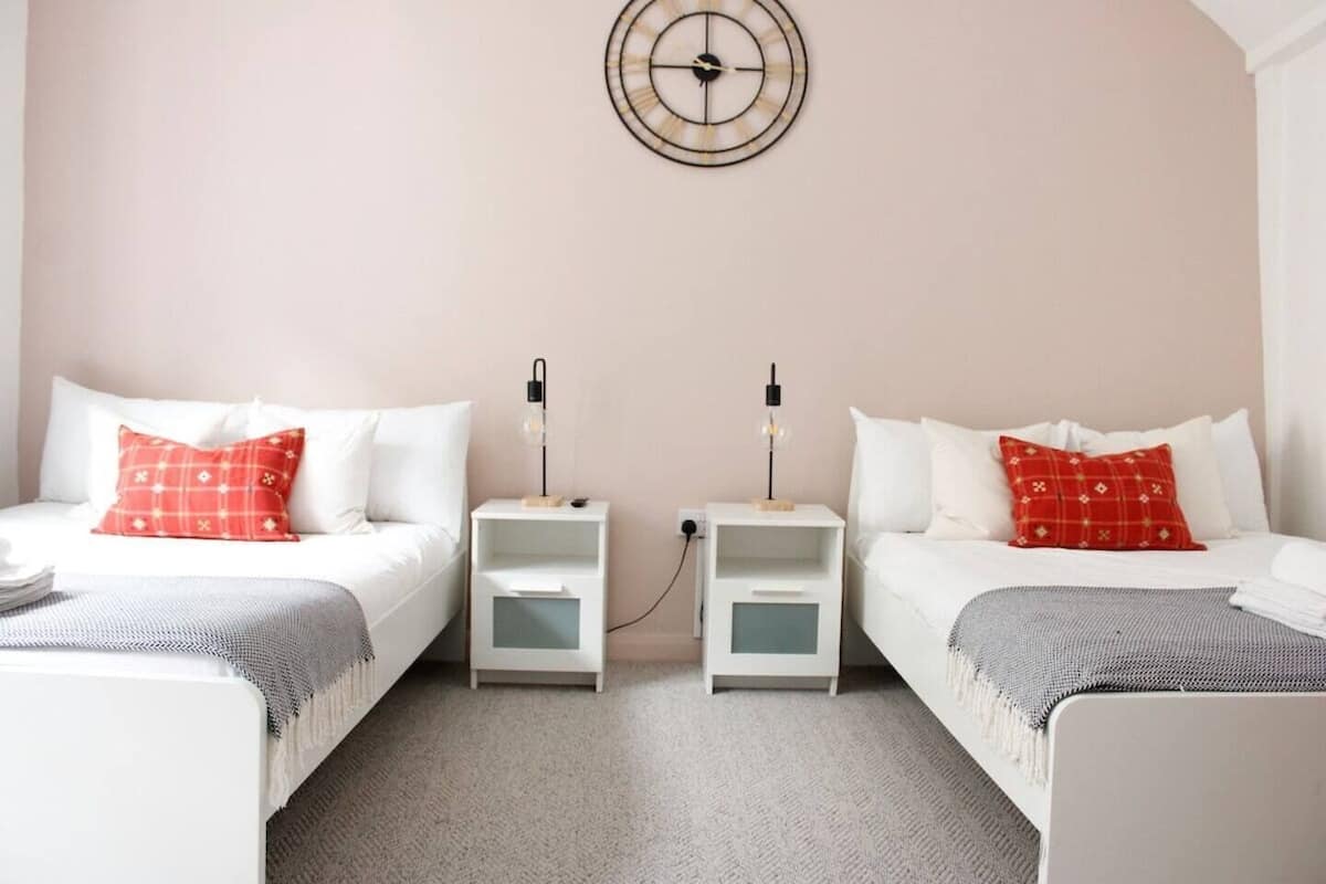 A twin room with two beds, each with a red pillow, a bedside table with a lamp, and a wall clock.