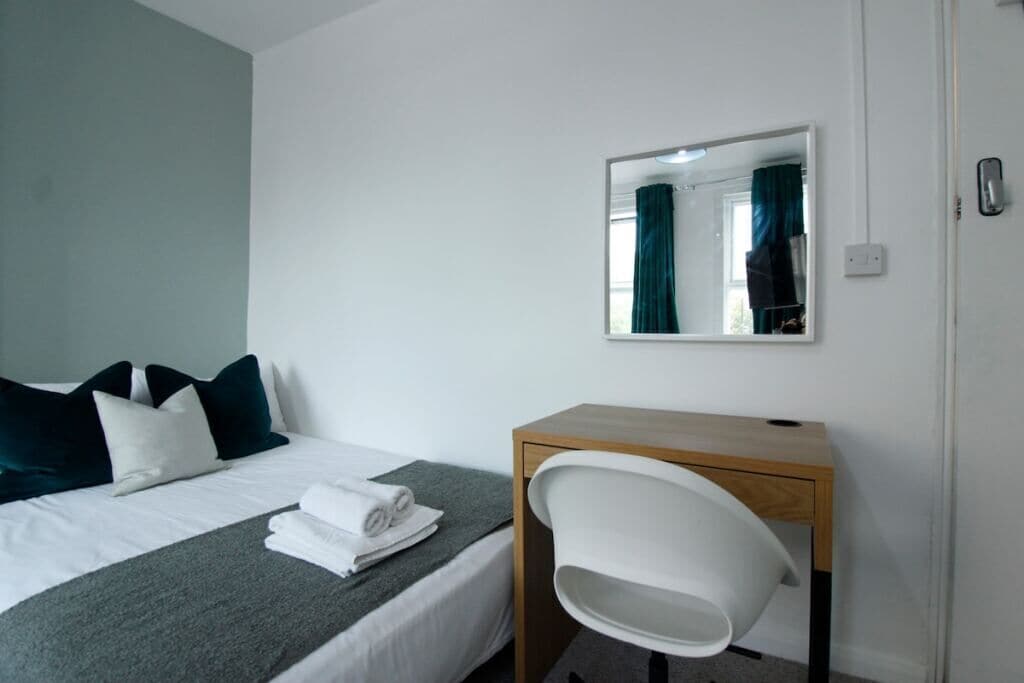 A hotel room with a bed, a desk, a chair, a mirror, and a window with curtains.