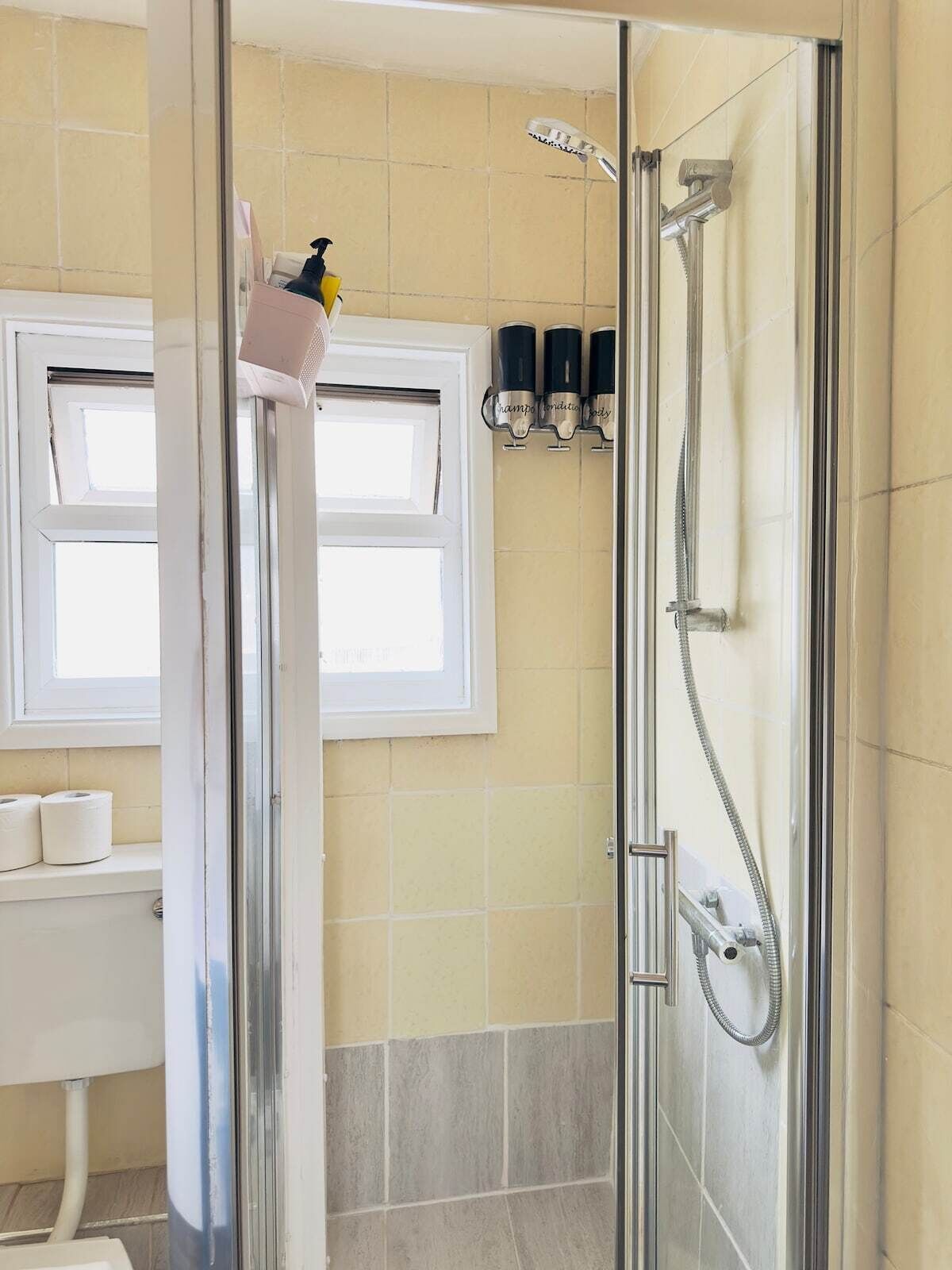 A bathroom with a glass shower enclosure, a window above the sink, and toiletries on the wall.