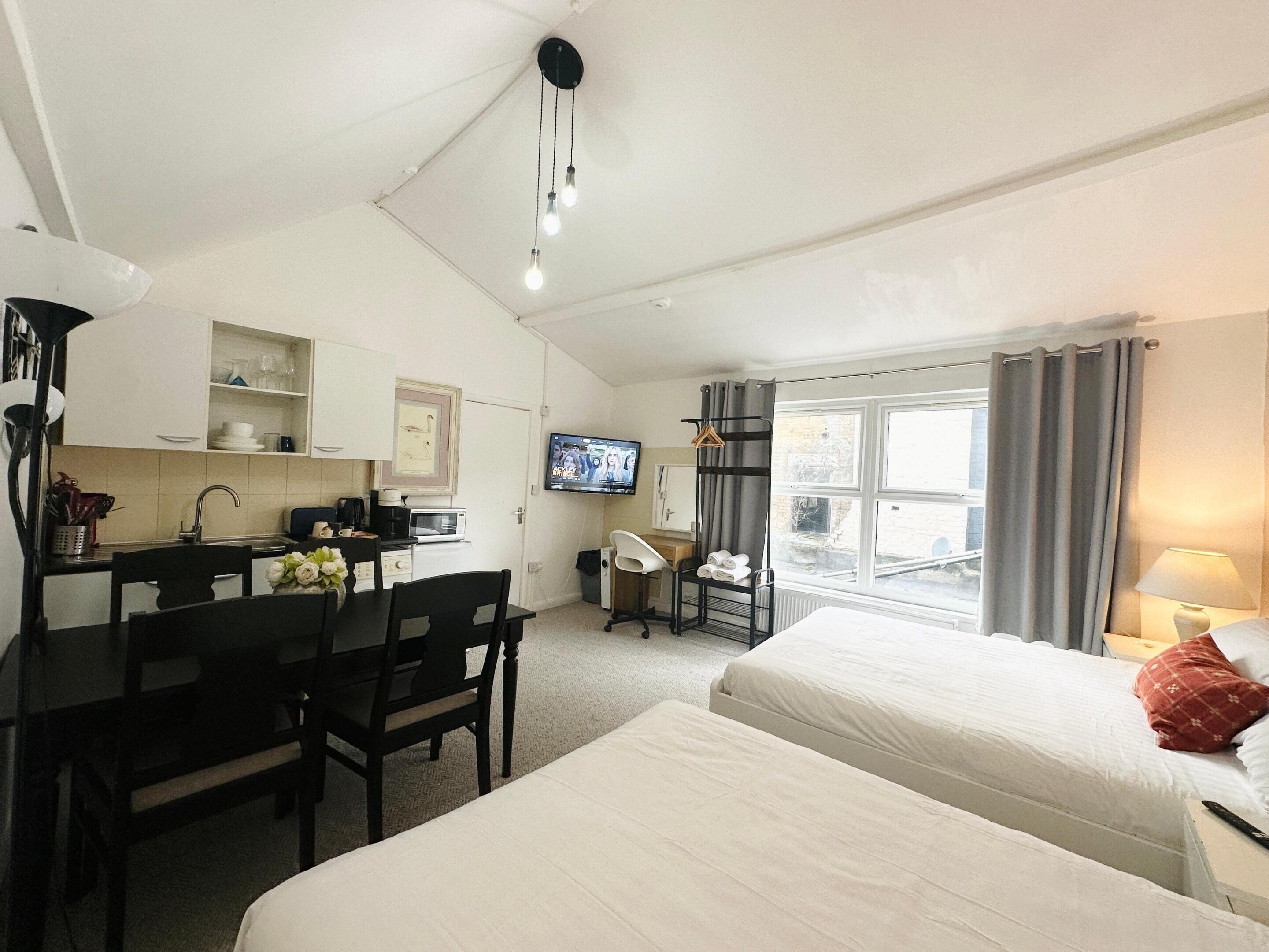 A compact hotel room with a kitchenette, a TV, a bed, and a window with curtains.