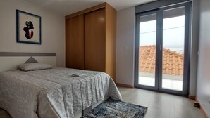 Et soveværelse med seng, et natbord, en lampe, et stort vindue og en skydedør til en balkon.