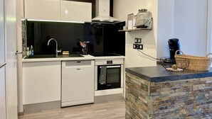 A modern kitchen with white cabinetry, a dishwasher, an oven, and a stone accent wall. - Proche du centre et plage, confort garanti (LA ROCHELLE)