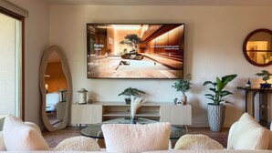 Ein modernes Wohnzimmer mit einem großen Flachbildfernseher, einem runden Couchtisch und einem Sofa. Zwei Beistelltische mit dekorativen Gegenständen und einer Topfpflanze ergänzen das Bild.