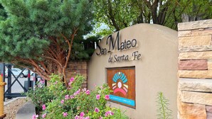 Schild mit der Aufschrift "San Mateo de Santa Fe" und einem Logo, umgeben von Grünpflanzen und einer Steinmauer.