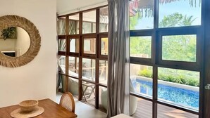 A room with a dining table, a large window with a view of a pool and greenery, and a woven light fixture. - Villa Uluwatu - Belle Vue Jungle, Villa 23 (Koh Samui)