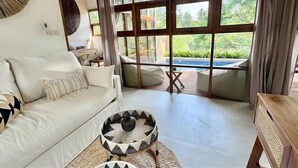 A modern living room with a white sofa, a round woven coffee table, and a view of a swimming pool through large windows. - Villa Uluwatu - Belle Vue Jungle, Villa 23 (Koh Samui)