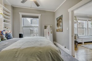 Une chambre avec un grand lit, une fenêtre donnant sur des immeubles, un ventilateur de plafond et un tableau accroché au mur.