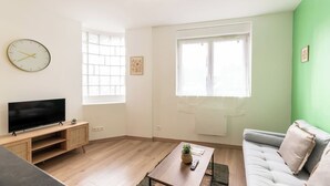 A living room with a flat-screen TV, wooden entertainment unit, a sofa, a coffee table, and a large window with blinds.