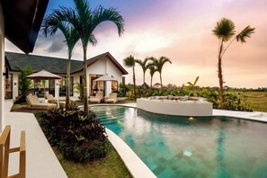 A modern poolside area with a curved pool, lounge chairs, and palm trees. - Villa Crystal Lagoon by BaliSuperHost (Bali)