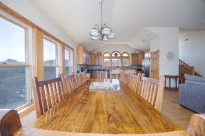 A spacious dining area with a long wooden table and chairs, a kitchen with wooden cabinets, and large windows offering a view. - 1825 - Off The Clock - Dog-Friendly Corolla Getaway w/ Pool (Corolla)